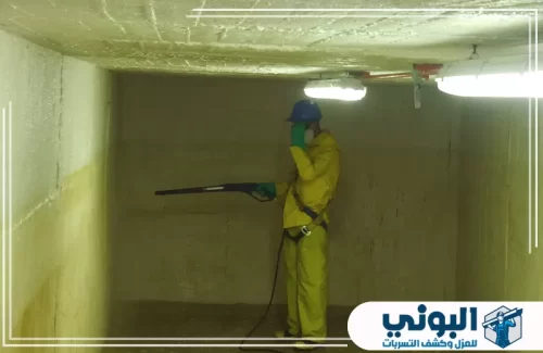 تنظيف الخزانات المعدنية بالدمام 0500805875 شركة البوني عامل ببدلة واقية صفراء وخوذة زرقاء يرش مادة بيضاء على الجدران والسقف داخل مساحة خرسانية مغلقة ومضاءة.